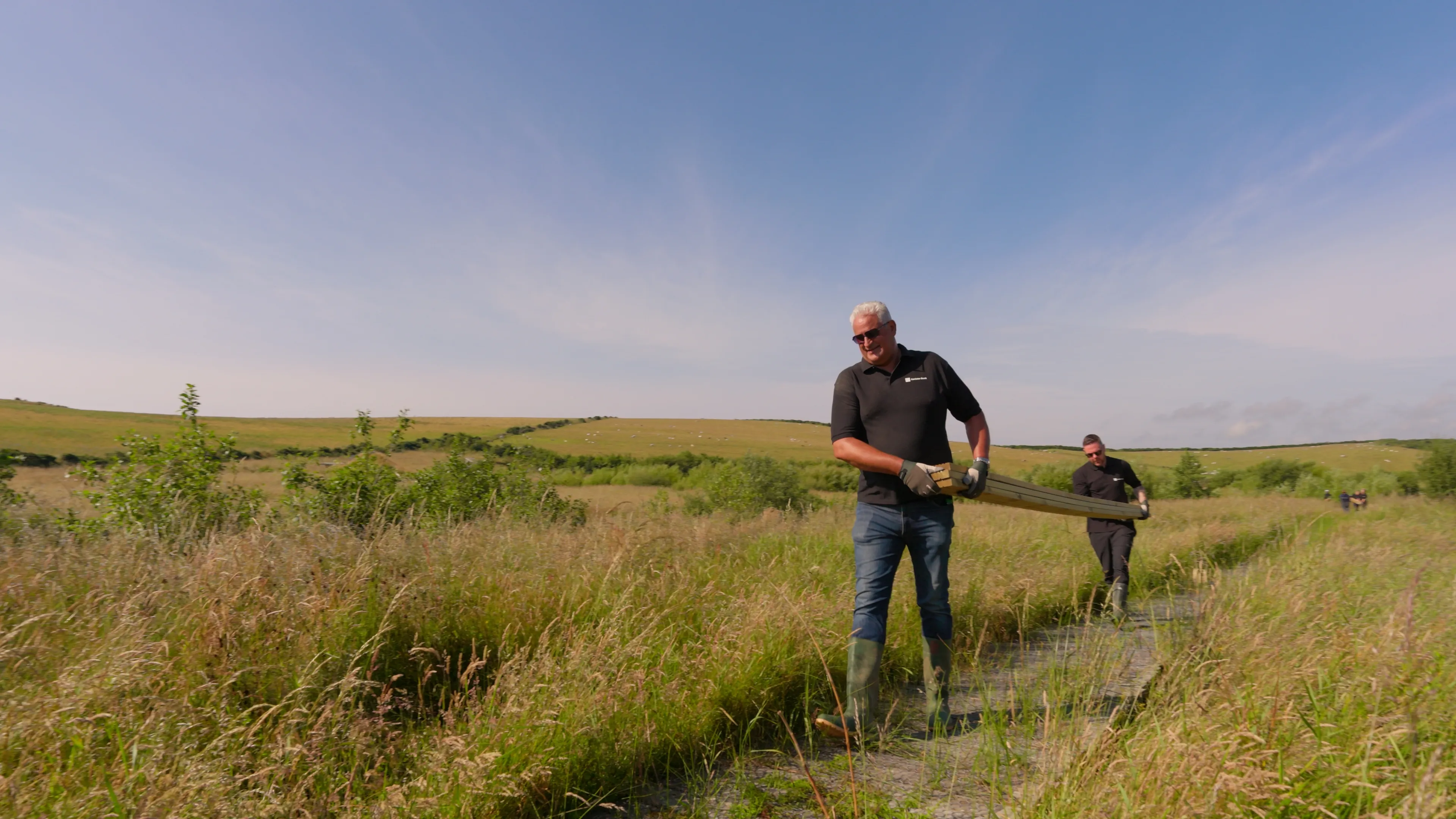 Two people carry a wooden log along a narrow path in a lush green field under a clear blue sky. The scene conveys teamwork and a serene rural setting.